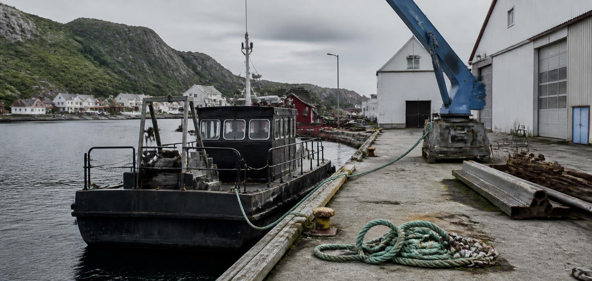 Arbeidsbåt fortøyd ved industrikai i et norsk kystmiljø med tauverk og kran