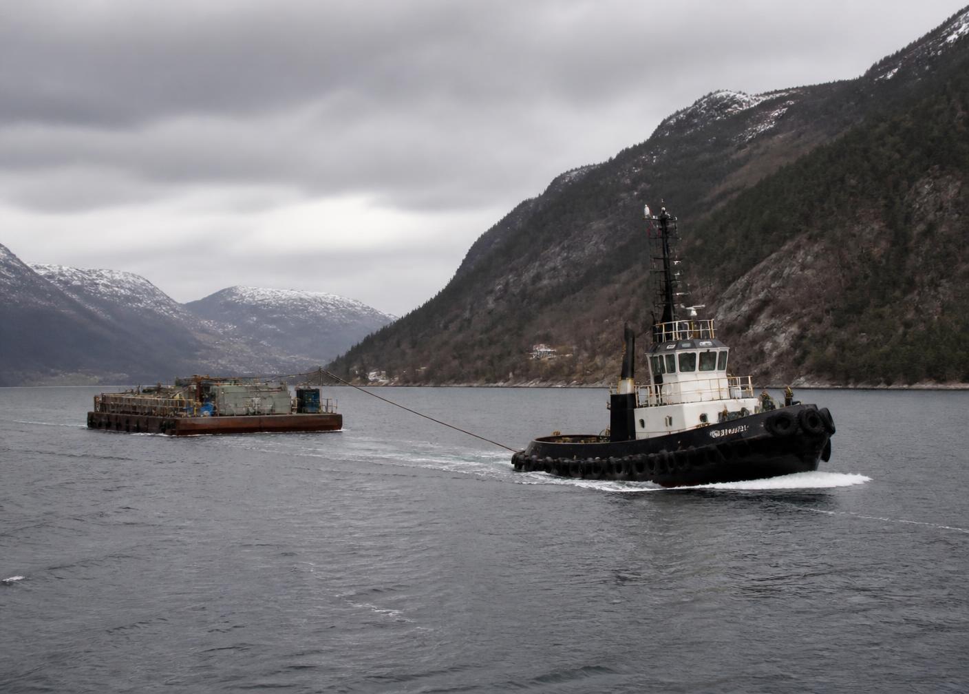 Slepebåt sleper en lekter gjennom en grå norsk fjord